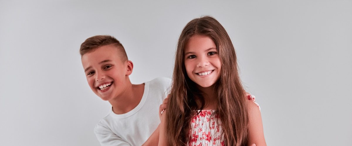 AdobeStock_267201078 Cute girl and boy on a gray background. The girl stands with arms crossed, and the boy looks out from behind her and smiles.